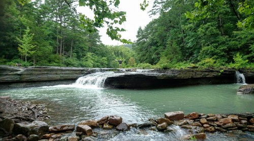 A serene landscape featuring a waterfall cascading into a calm green pool, surrounded by lush trees and rocky terrain.