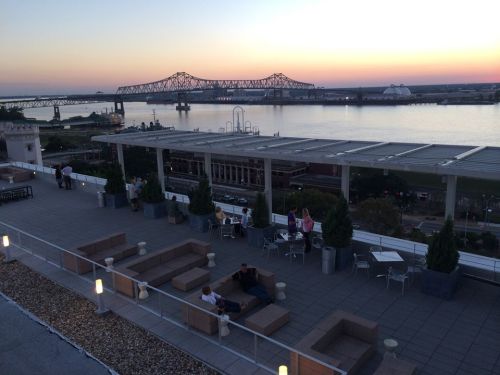 Rooftop view at sunset overlooking a river, with seating areas and a bridge in the background.