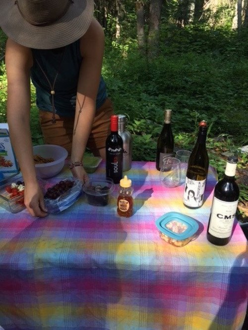 A person prepares food on a colorful tablecloth in a forest, surrounded by wine bottles and snacks.