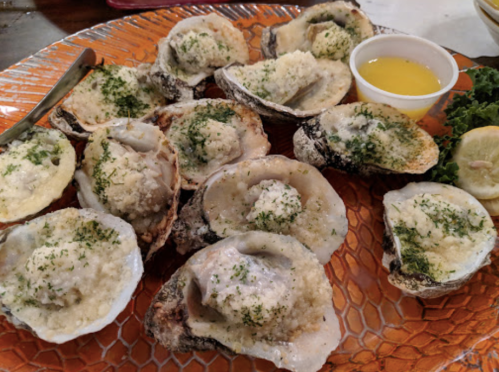 A platter of baked oysters topped with breadcrumbs and herbs, served with lemon and a small cup of dipping sauce.
