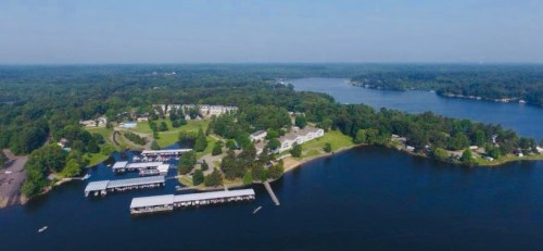 Aerial view of a serene lake surrounded by green trees and a marina with boats, under a clear blue sky.