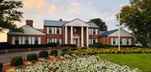 A large brick house with white columns, surrounded by manicured lawns and colorful flower beds under a cloudy sky.