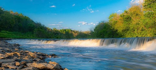 A serene river flows over a waterfall, surrounded by lush greenery and a clear blue sky with scattered clouds.