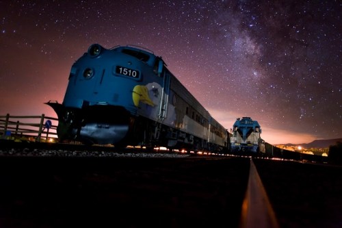 A blue train under a starry night sky, with the Milky Way visible in the background.