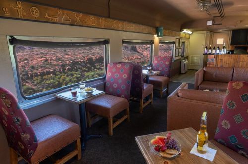 Interior of a train car with cozy seating, tables, and large windows showcasing a scenic view outside.