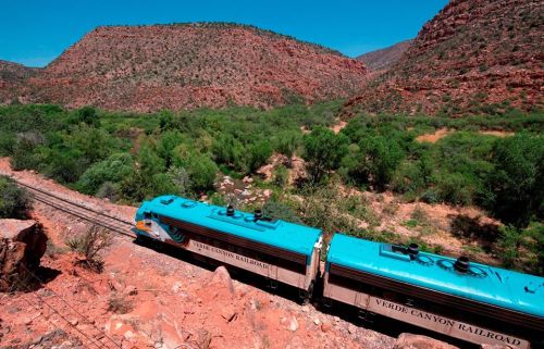 A turquoise train travels along tracks through a lush green valley surrounded by red rock mountains.