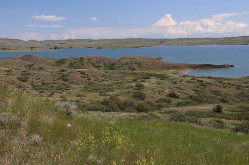A serene landscape featuring rolling hills, green vegetation, and a calm blue lake under a partly cloudy sky.