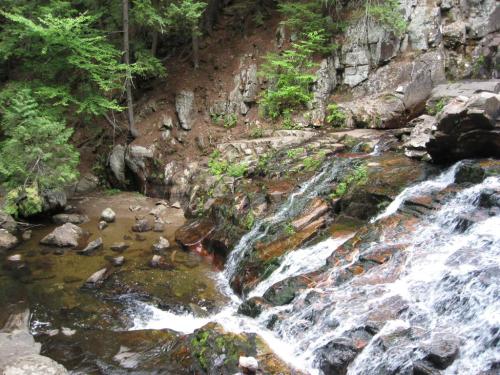 A serene waterfall cascades over rocks into a clear stream, surrounded by lush greenery and rugged terrain.