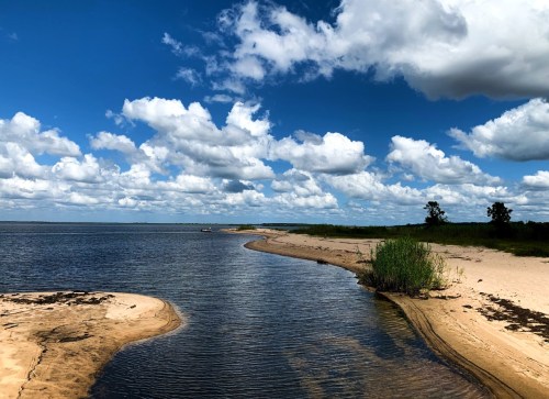 A serene landscape featuring a winding river, sandy banks, and fluffy clouds against a bright blue sky.