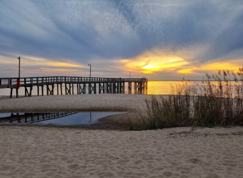 A serene beach scene at sunset, featuring a wooden pier extending over calm waters and soft sand in the foreground.
