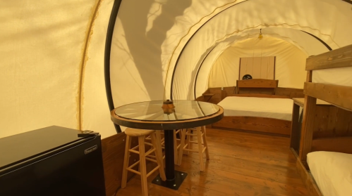 Cozy interior of a glamping tent featuring a bed, table with stools, and a small fridge. Warm, inviting atmosphere.