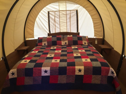 Cozy interior of a covered wagon featuring a large bed with a patchwork quilt and two side tables.