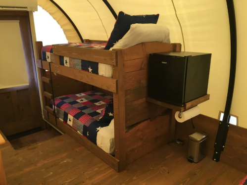 Cozy interior of a glamping tent featuring a wooden bunk bed, colorful quilts, and a mini fridge.