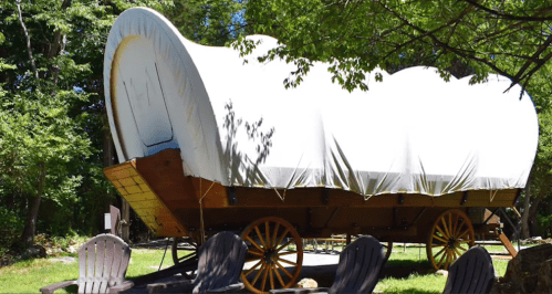 A covered wagon on a grassy area surrounded by trees, with wooden chairs in front.