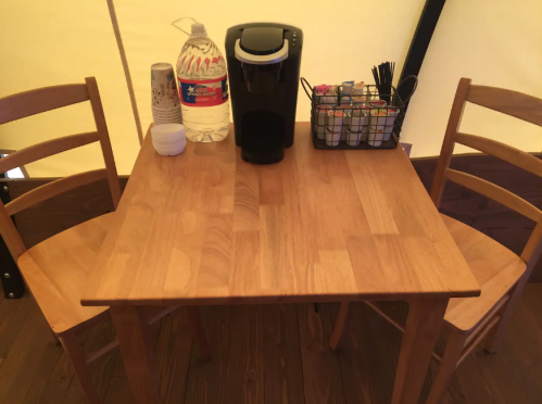 A wooden table with two chairs, a water bottle, a coffee maker, and a basket of condiments inside a tent.