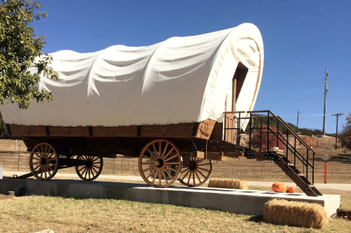 A covered wagon on a platform, with wooden wheels, set against a clear blue sky and grassy field.