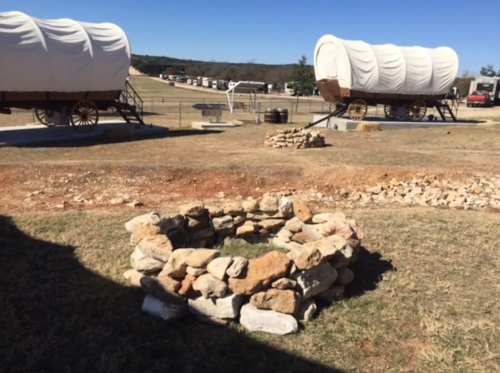 Two covered wagons on a grassy field, with a stone fire pit in the foreground and a clear blue sky above.