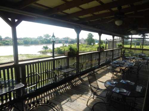 A riverside restaurant patio with empty tables, overlooking a calm river and green landscape under a wooden roof.