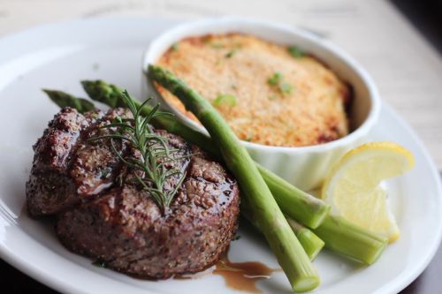 A plate featuring a grilled steak with rosemary, asparagus, a lemon wedge, and a side of creamy baked dish.