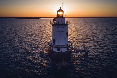A lighthouse stands on rocky shores at sunset, casting a warm glow over the calm waters.