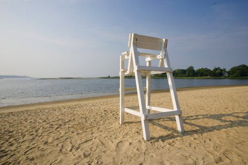 A white lifeguard chair on a sandy beach overlooking a calm water body under a clear sky.