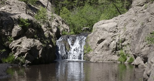 A serene waterfall cascades into a calm pool, surrounded by rocky cliffs and lush greenery.