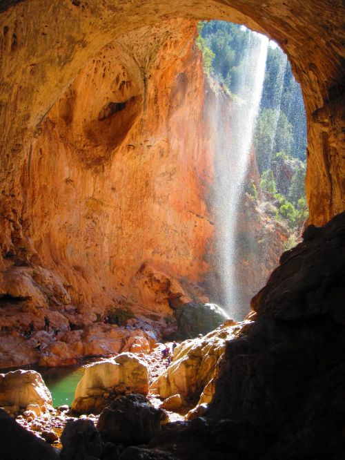 A stunning cave with a waterfall cascading down orange rock formations, surrounded by lush greenery and a serene pool below.