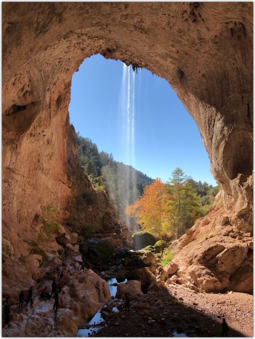 A stunning cave opening with a waterfall, surrounded by trees and rocky terrain under a clear blue sky.