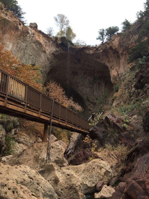 A wooden bridge spans rocky terrain, leading to a large cave opening surrounded by autumn foliage and steep cliffs.