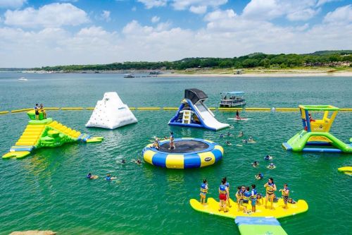 A vibrant water park scene with inflatable structures, people swimming, and a clear blue sky over a lake.