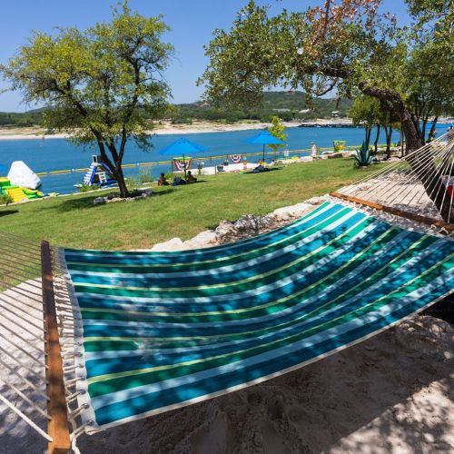 A colorful hammock sways between trees, overlooking a serene lake and grassy area with recreational activities.