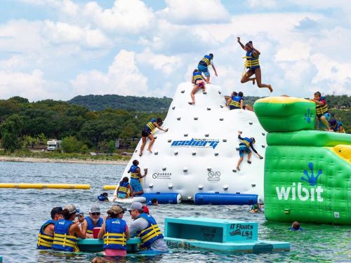 A group of people in life jackets enjoying a water park with inflatable structures and a climbing wall on a lake.