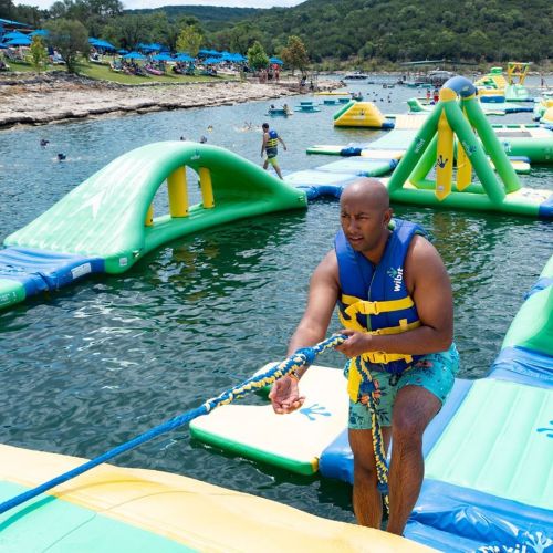 A man in a life vest pulls a rope on a floating obstacle course in a lake, surrounded by people enjoying the water.