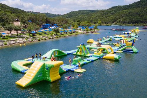 A vibrant inflatable water park with slides and obstacles on a lake, surrounded by green hills and blue skies.