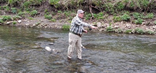 A person stands in a stream, casting a fishing line while wearing a plaid shirt and fishing gear.