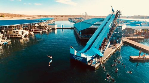 A water slide at a marina with people sliding down and swimming in the water, surrounded by boats and blue skies.