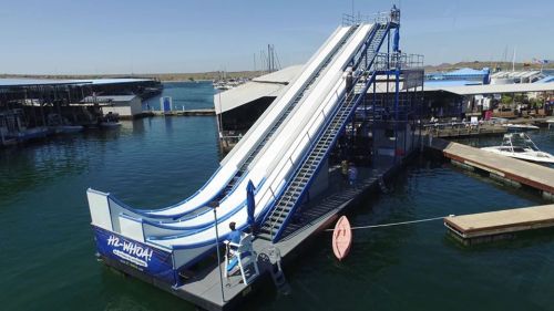 A large water slide at a marina, with boats docked nearby and a clear blue sky above.