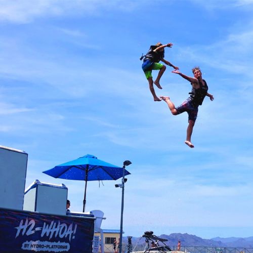 Two people jumping off a platform into the water, with a blue sky and mountains in the background.