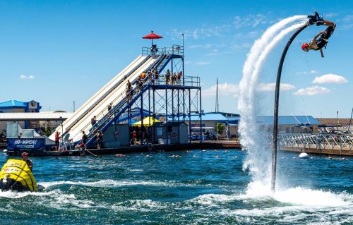 A person performs a stunt on a water jetpack near a waterslide and spectators at a sunny water park.
