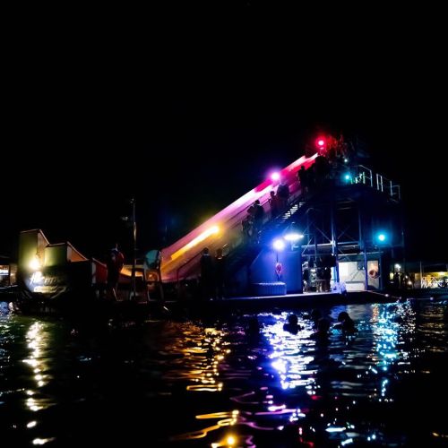 A brightly lit water slide at night, with people swimming nearby and colorful lights reflecting on the water.