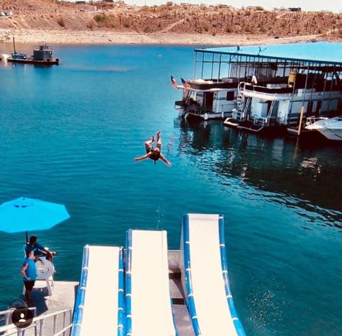 A person jumps off a diving board into a lake, with houseboats and a sunny shoreline in the background.