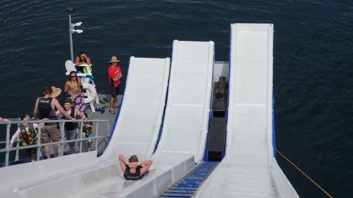 A view of three water slides leading into a body of water, with people waiting and relaxing nearby.