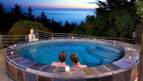 A couple relaxes in a hot tub overlooking a serene ocean view at sunset, surrounded by candles and greenery.