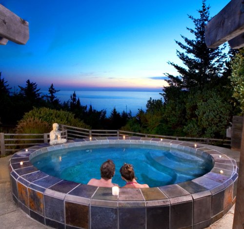 A couple relaxes in a hot tub at sunset, overlooking a serene ocean view surrounded by trees.