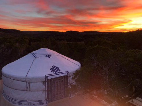 A yurt sits on a hillside under a vibrant sunset, surrounded by trees and rolling hills.
