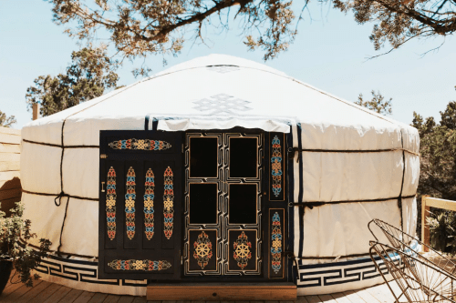 A traditional yurt with ornate wooden doors, set against a clear blue sky and surrounded by trees.