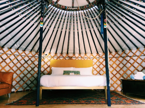 Cozy yurt interior featuring a large bed, decorative pillows, and wooden lattice walls. Warm, inviting atmosphere.