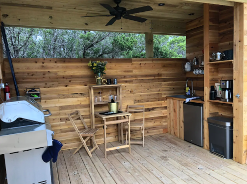 Cozy outdoor kitchen with wooden walls, a small table, chairs, and appliances, surrounded by greenery.