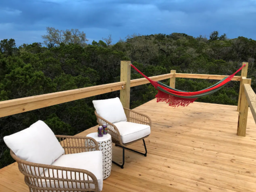 A wooden deck with two chairs, a small table, and a colorful hammock, surrounded by trees under a cloudy sky.