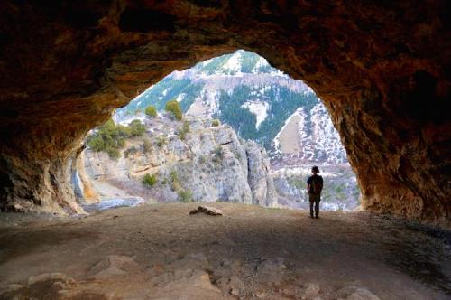 A person stands inside a cave, looking out at a mountainous landscape through the cave's opening.